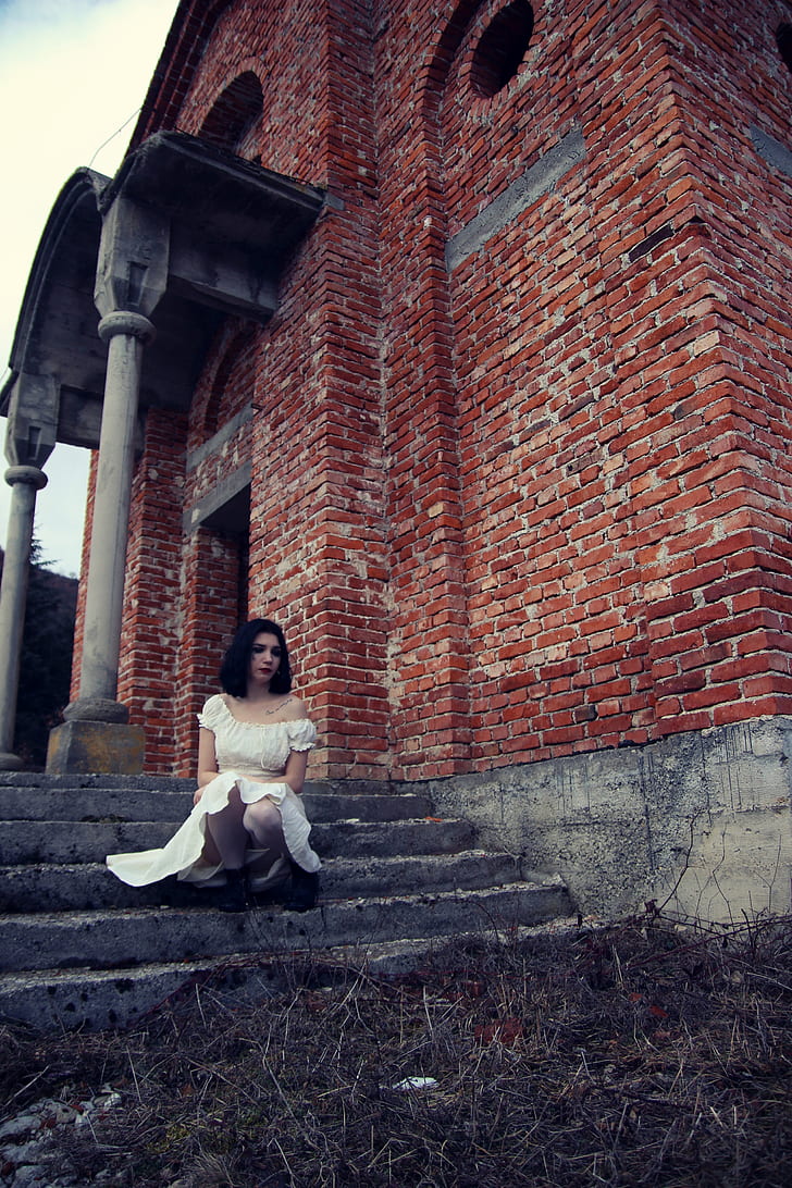 photo of woman wearing off-shoulder dress sitting on stair of brown concrete building