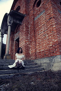 photo of woman wearing off-shoulder dress sitting on stair of brown concrete building