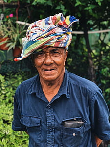 man wearing blue button-up collared t-shirt and multicolored headdress standing near green leaf plants during daytime